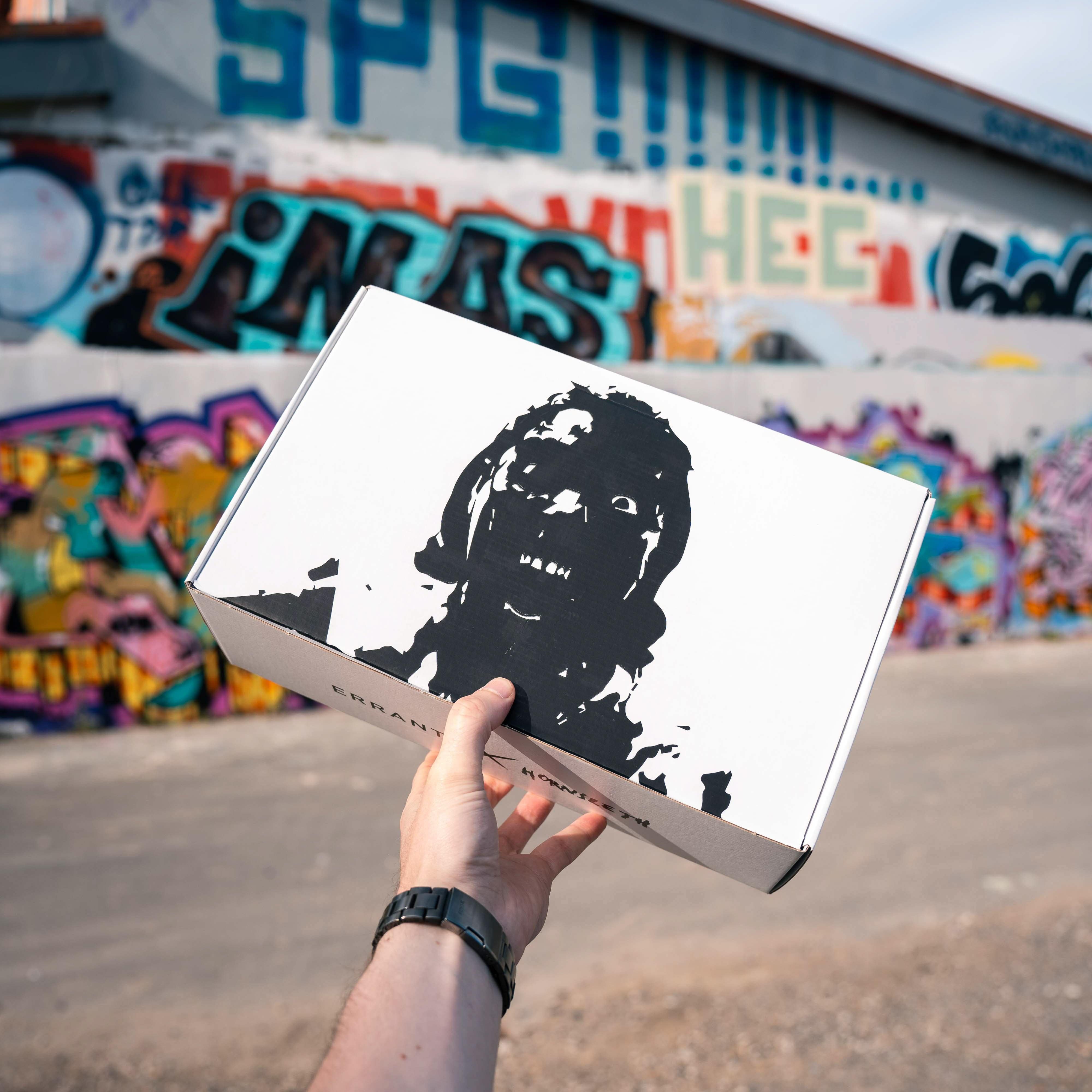 En person holder en hvid kasse med et sort ansigtstryk foran en farverig graffitimur.