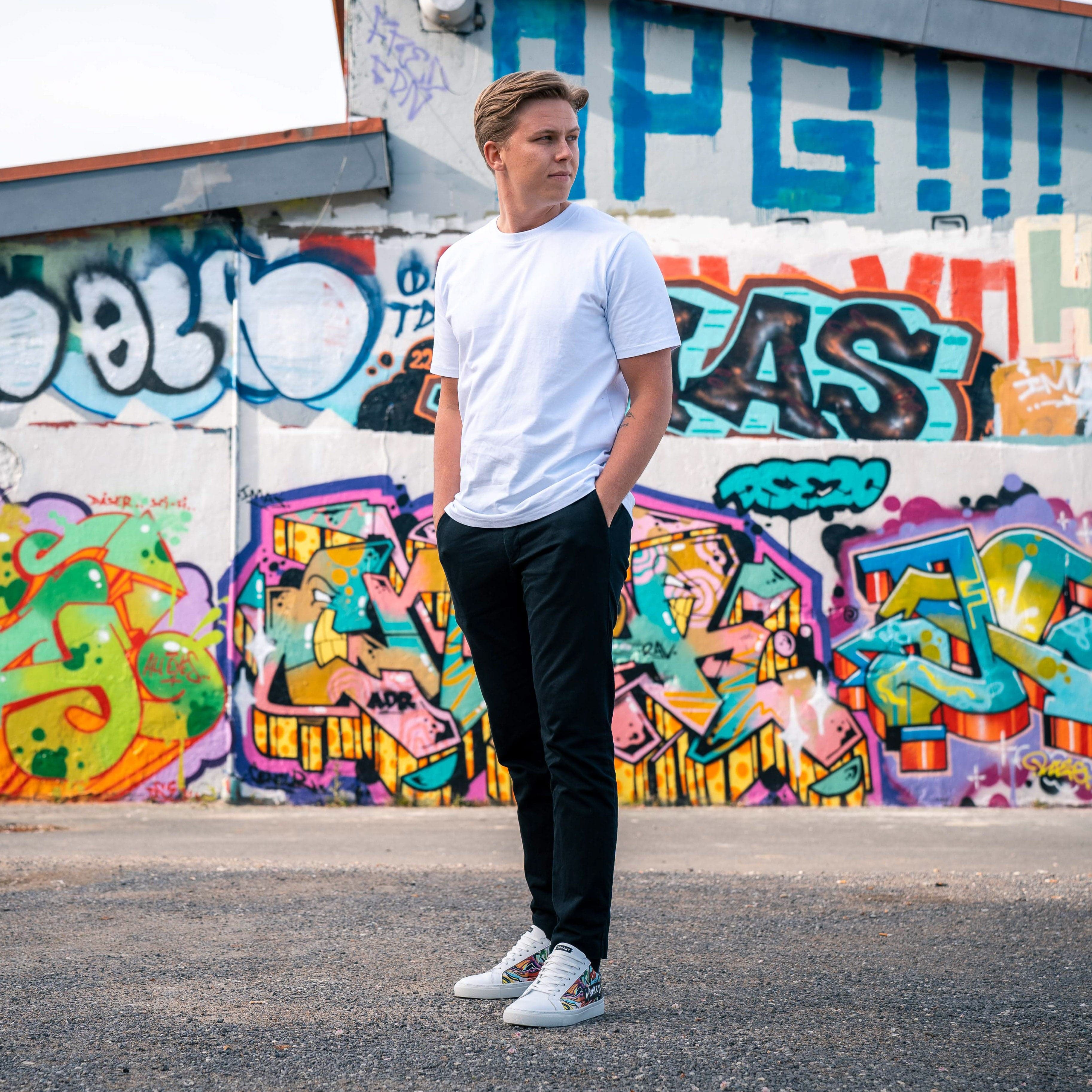 En mand iført en hvid t-shirt og sorte bukser står foran en farverig graffitimur.