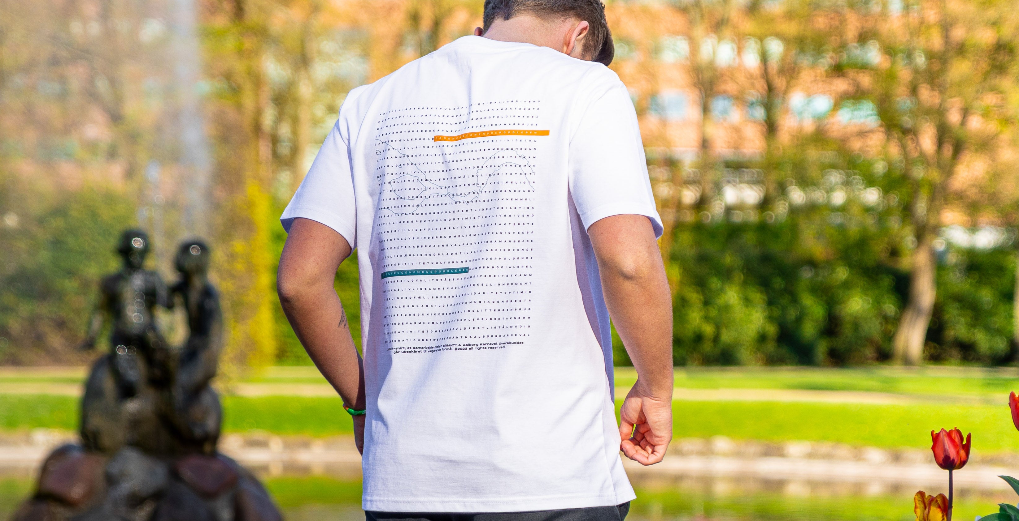 En person iført en hvid t-shirt med et grafisk design på ryggen, står i en park.