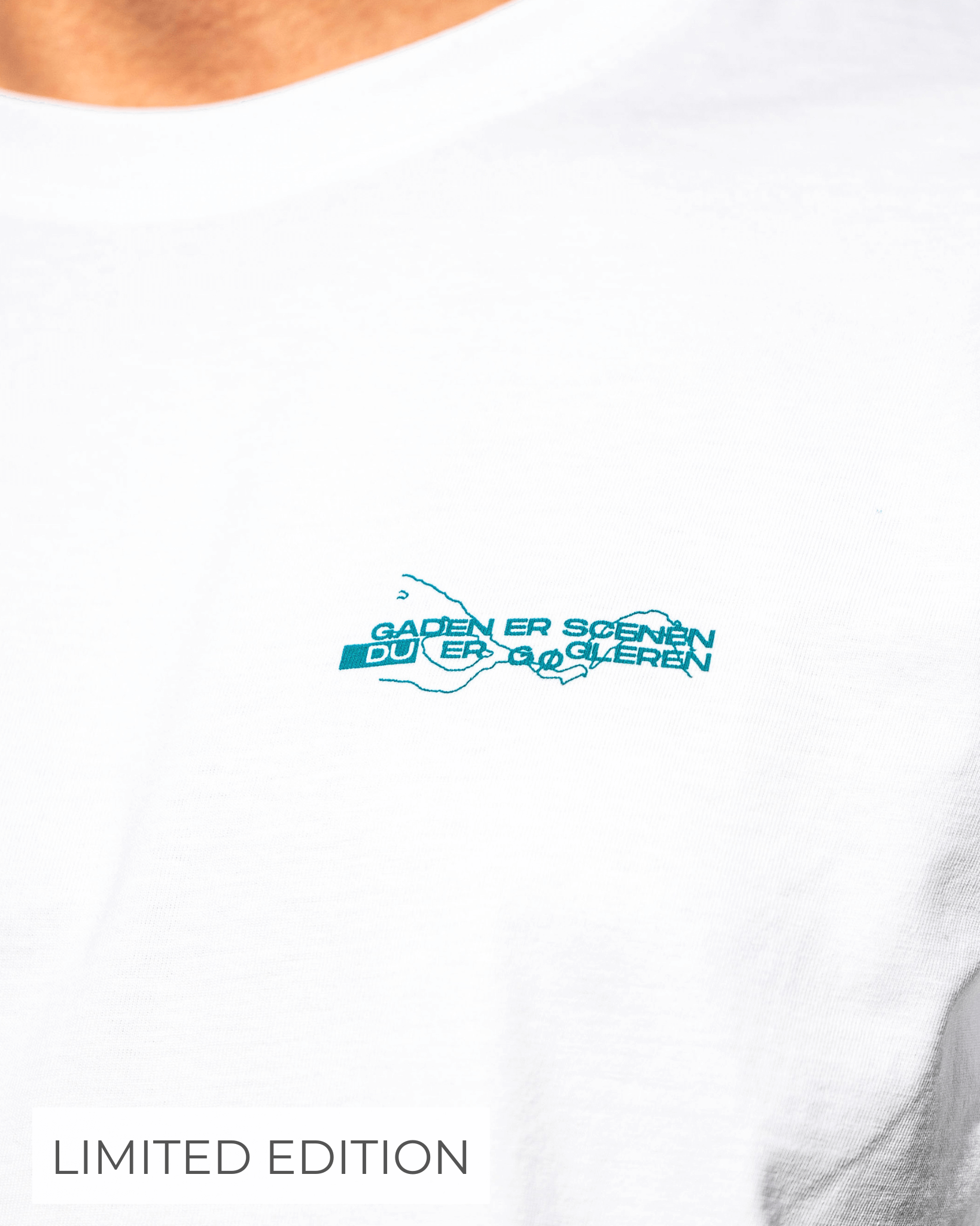 Billedet viser en hvid t-shirt med en lille blå tekst og grafik på brystet, hvor der står "GADEN ER SCENEN DU ER SKUESPILLEREN". Nederst til venstre står der "LIMITED EDITION".