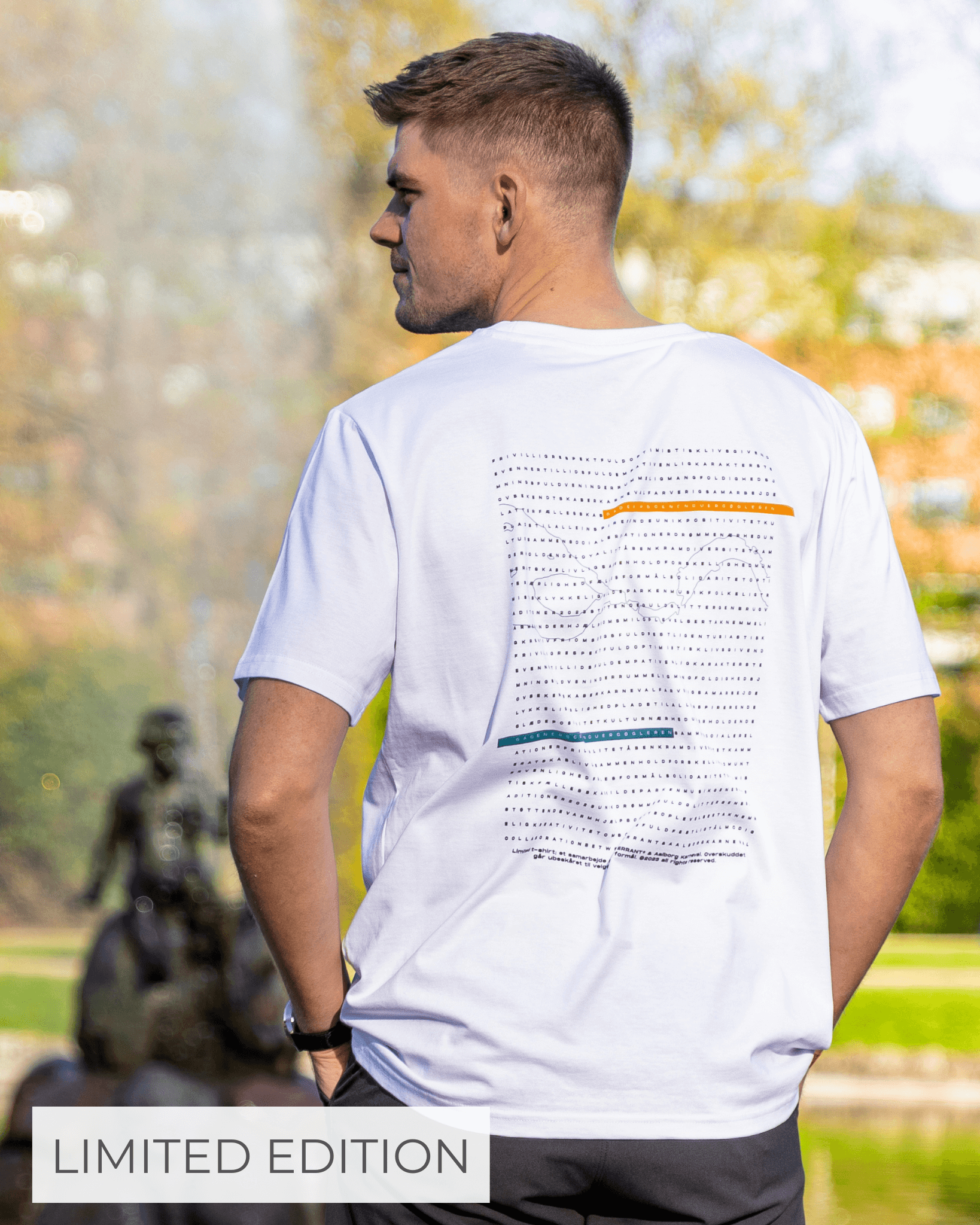 En mand står med ryggen til iført en hvid T-shirt med et grafisk mønster på ryggen. I baggrunden ses en sløret park med en statue. Teksten "LIMITED EDITION" er synlig nederst i billedet.