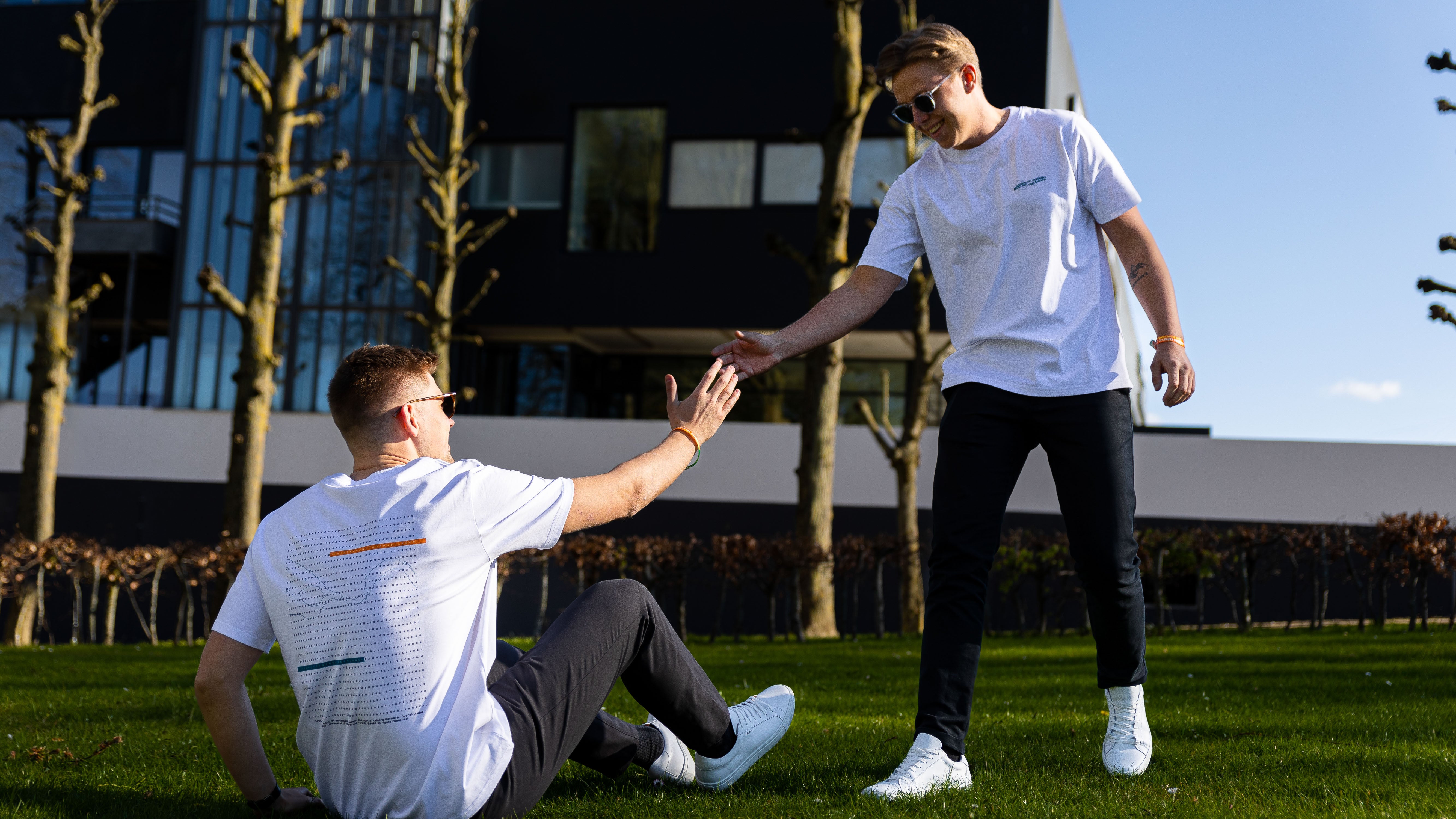 To mænd iført hvide t-shirts, der giver hinanden high-five på en græsplæne foran en moderne bygning.