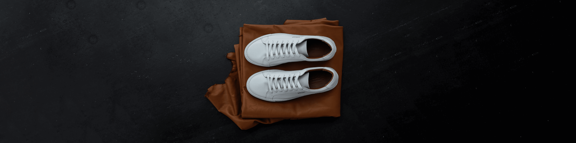 Et par hvide sneakers ovenpå en brun jakke på en sort overflade.