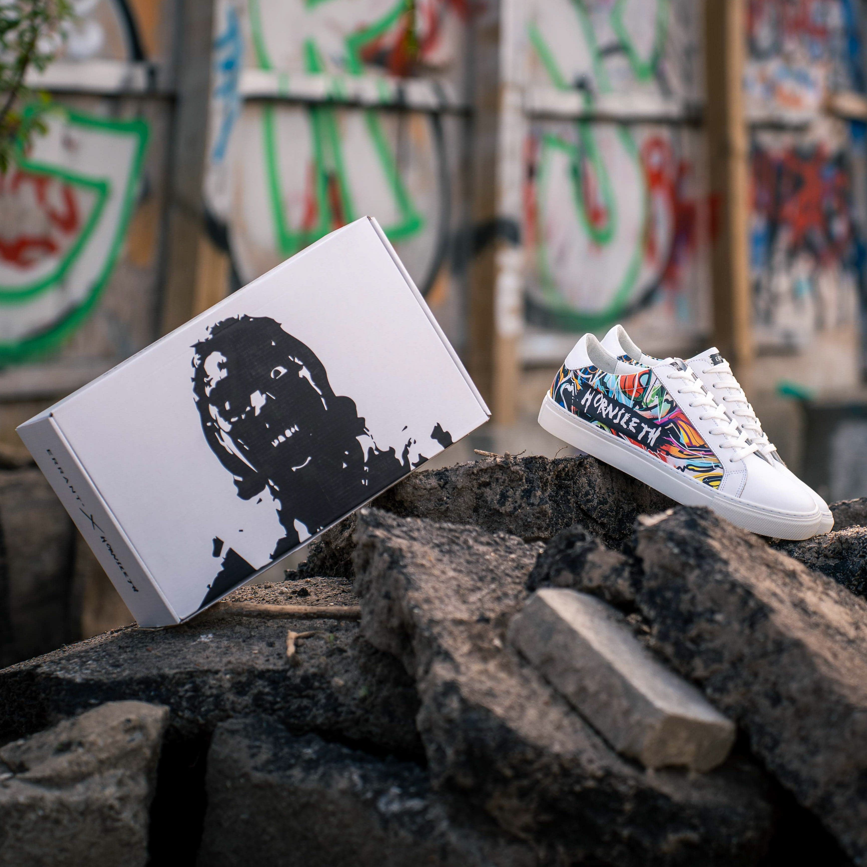 Et par farverige sneakers og deres æske er placeret på nogle betonblokke foran en væg med graffiti.
