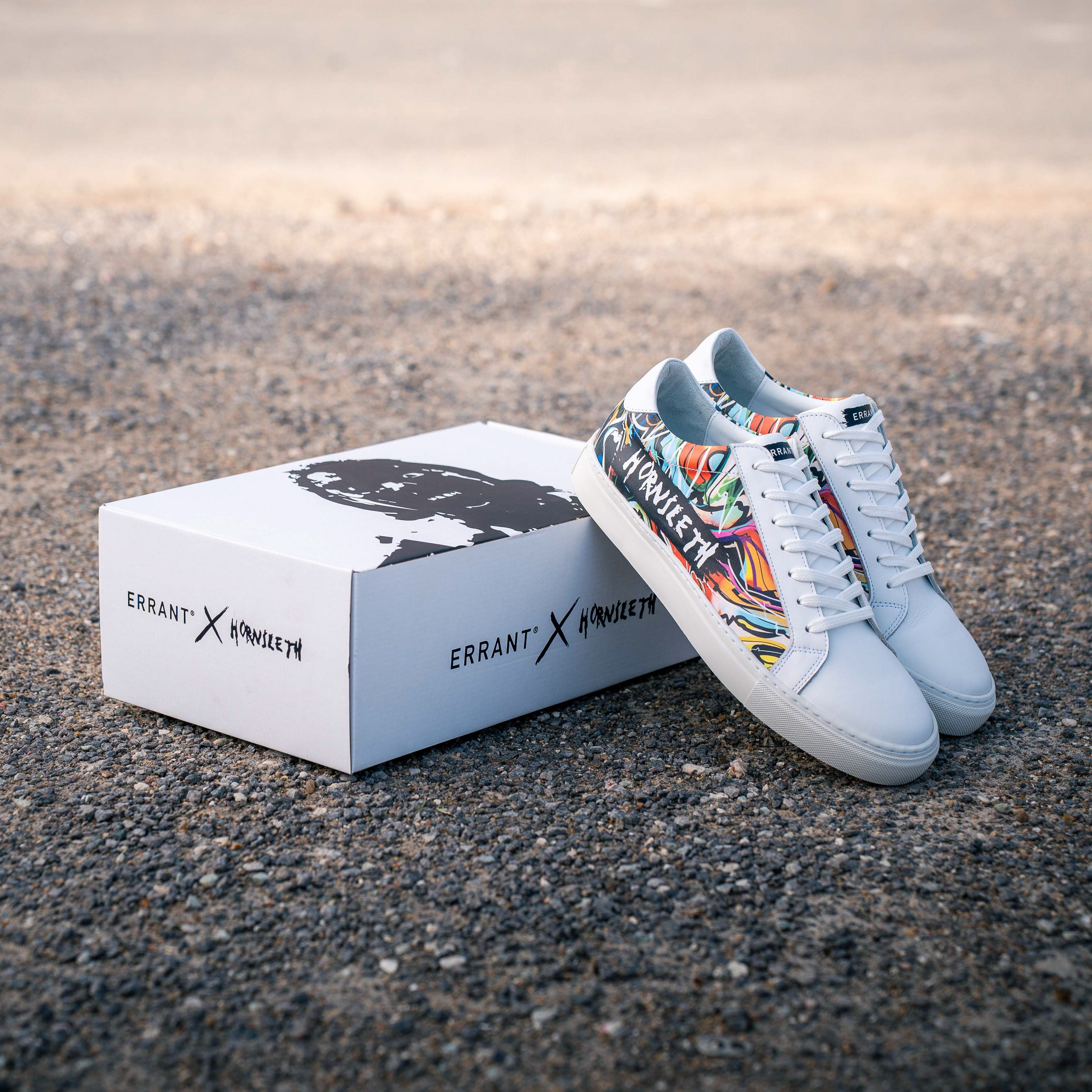 Billedet viser et par hvide sneakers med farverige detaljer ved siden af en hvid skoæske med teksten "ERRANT X MANVSLTH".