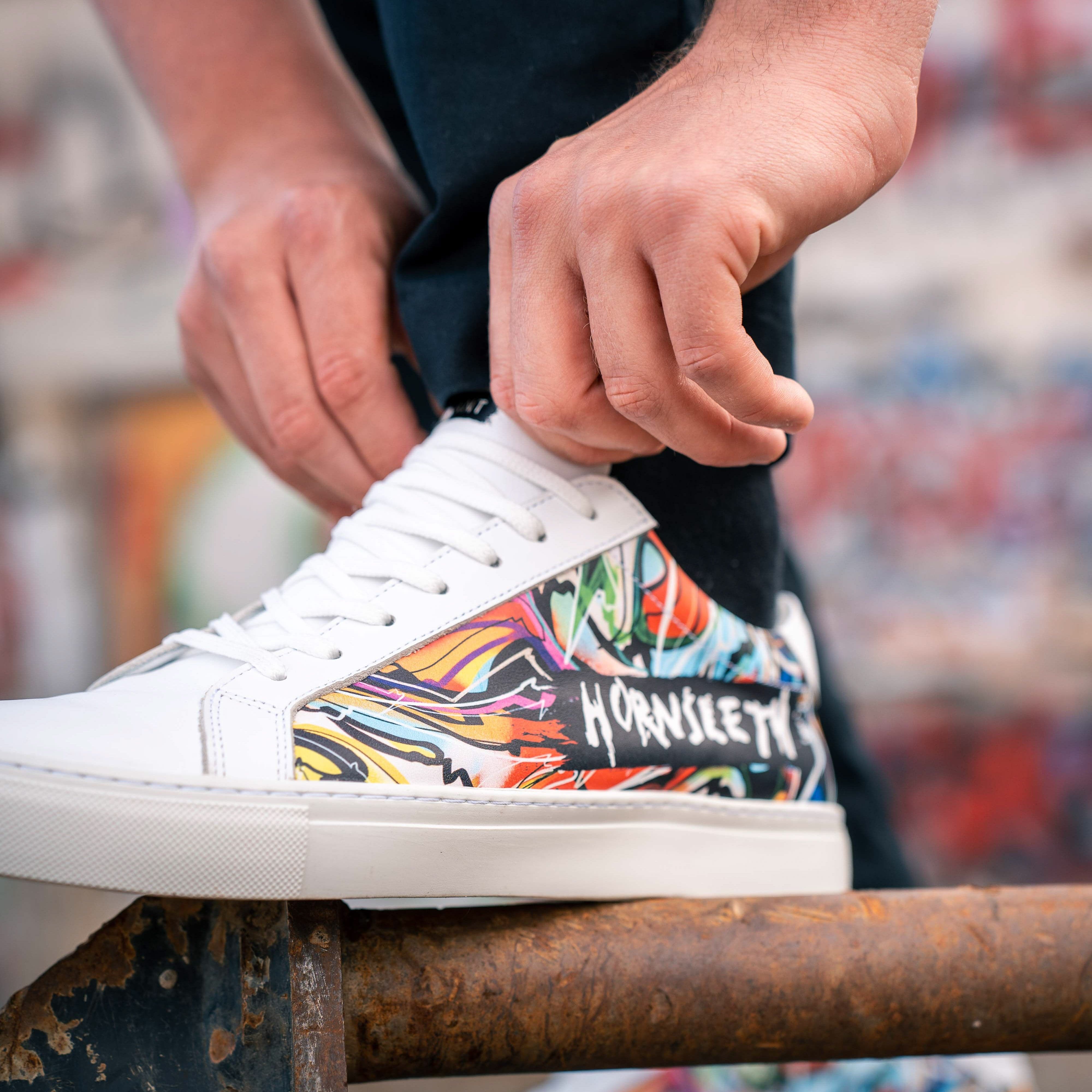 En person snører et par hvide sneakers med farverigt, grafisk design og teksten "HØRNSETH" på siden.