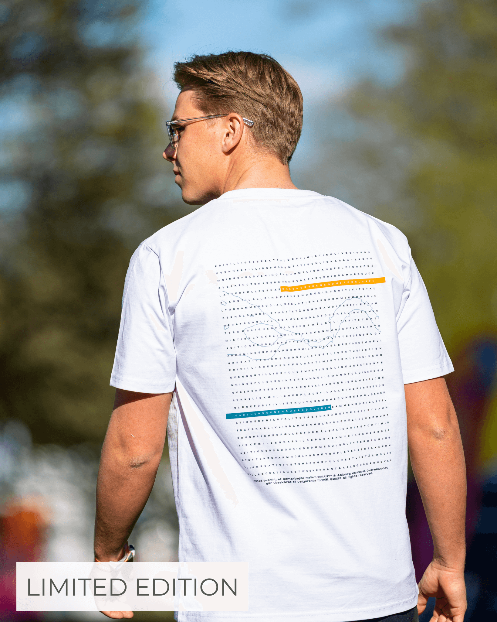 En mand iført en hvid T-shirt med et grafisk design på ryggen står udendørs. Teksten "LIMITED EDITION" er synlig i nederste venstre hjørne.