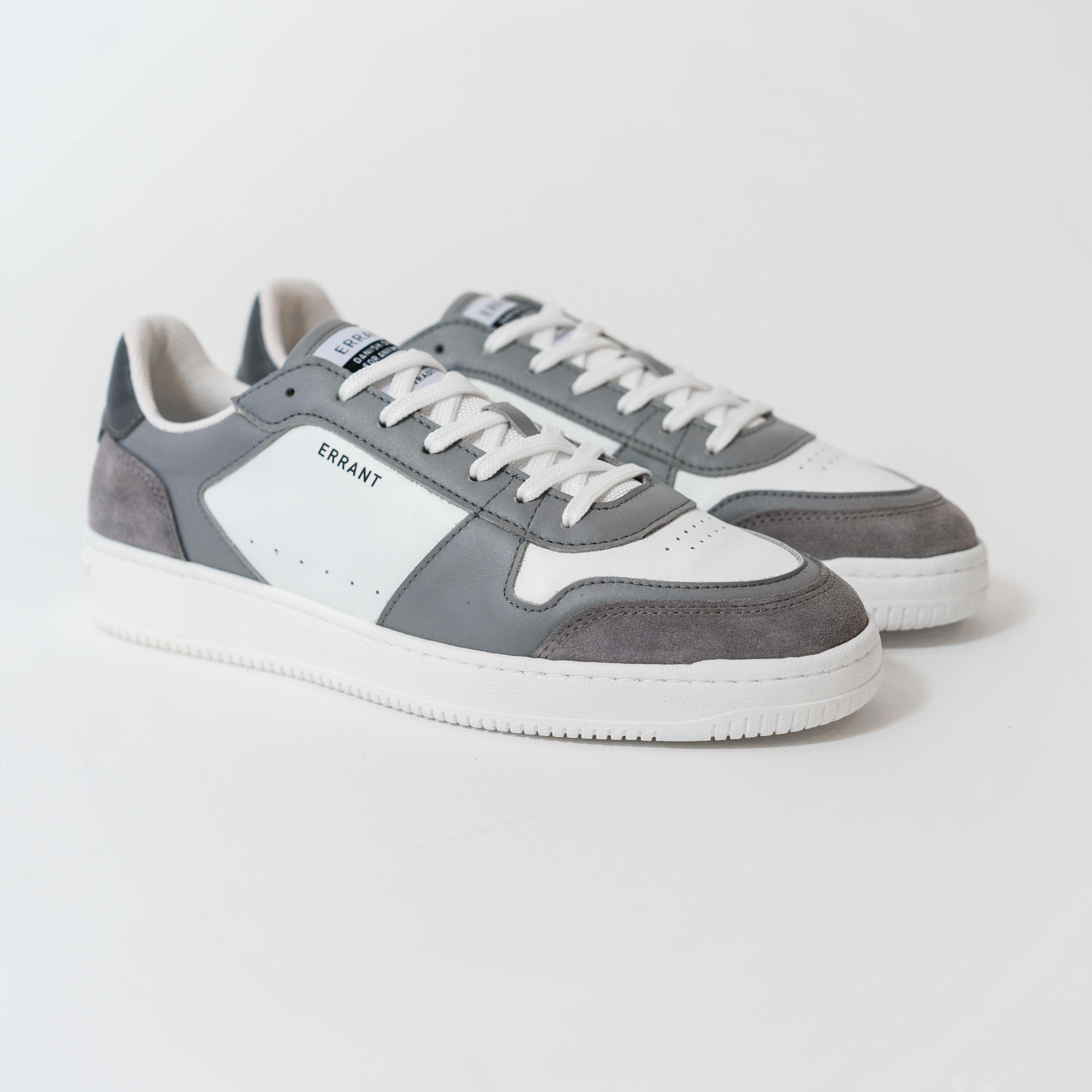 Billedet viser et par grå og hvide sneakers med snørebånd og en hvid sål.