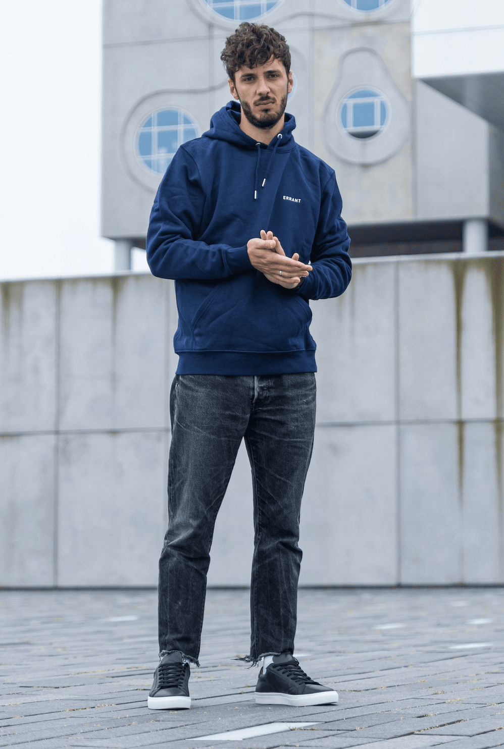 Mand i blå hættetrøje og sorte jeans står foran moderne bygning.